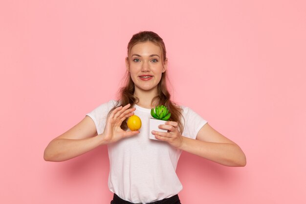 Vue de face de la jeune femme en t-shirt blanc tenant une petite plante de citron frais sur un mur rose clair