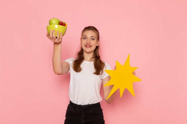 Vue de face de la jeune femme en t-shirt blanc tenant la plaque avec des fruits et grand panneau jaune souriant sur mur rose