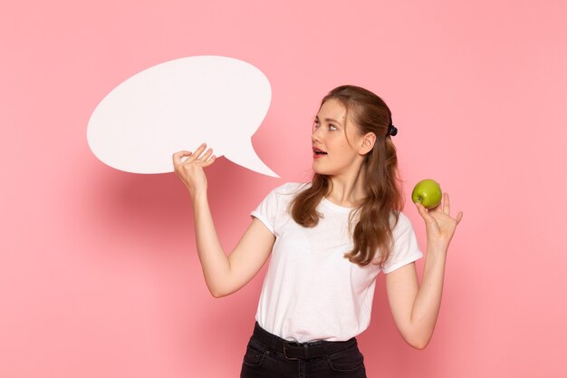 Vue de face de la jeune femme en t-shirt blanc tenant une pomme verte fraîche et grand panneau blanc sur un mur rose clair