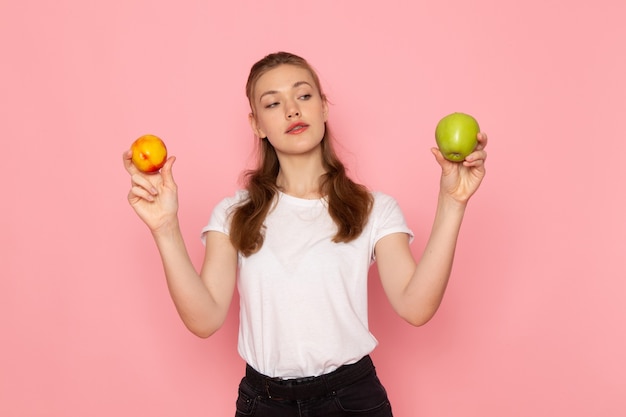 Vue de face de la jeune femme en t-shirt blanc tenant pomme verte fraîche avec pêche sur mur rose clair