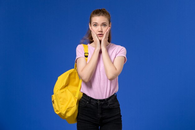 Vue de face de la jeune femme en t-shirt rose portant un sac à dos jaune posant sur le mur bleu