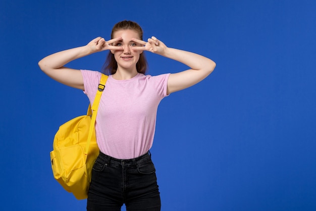 Vue de face de la jeune femme en t-shirt rose portant un sac à dos jaune souriant juste sur le mur bleu clair