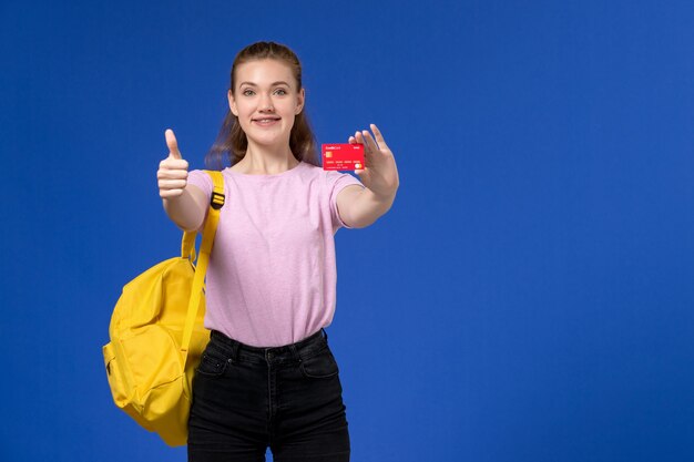 Vue de face de la jeune femme en t-shirt rose portant un sac à dos jaune tenant une carte rouge en plastique souriant sur mur bleu