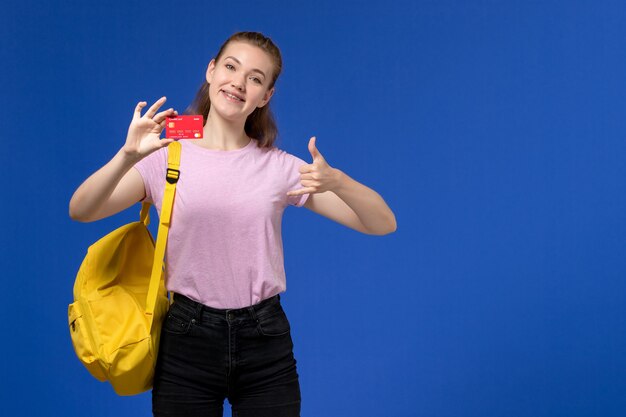 Vue de face de la jeune femme en t-shirt rose portant un sac à dos jaune tenant une carte rouge en plastique avec sourire sur le mur bleu