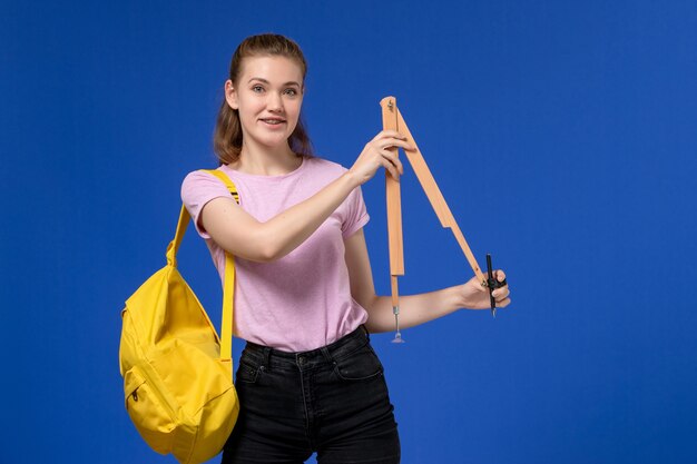 Vue de face de la jeune femme en t-shirt rose portant un sac à dos jaune tenant une figure en bois souriant sur un mur bleu