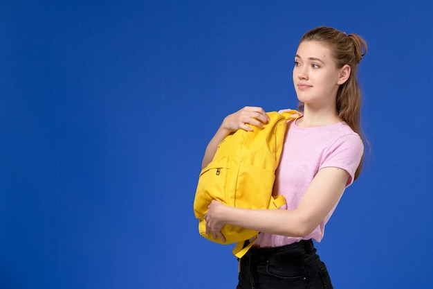 Vue de face de la jeune femme en t-shirt rose tenant un sac à dos jaune sur le mur bleu