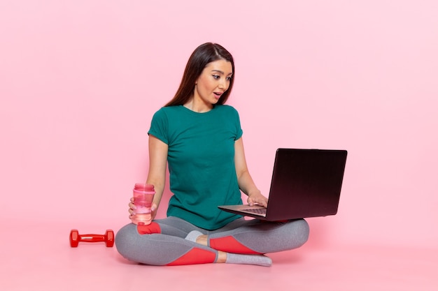 Vue de face jeune femme en t-shirt vert à l'aide d'un ordinateur portable sur le mur rose exercice taille exercice beauté slim sport féminin