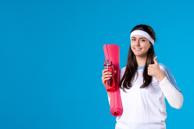 Vue de face jeune femme avec tapis pour les exercices et bouteille d'eau sur le mur bleu