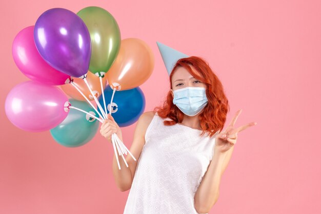 Vue de face jeune femme tenant des ballons colorés en masque sur plancher rose xmas color virus pandemic covid- party