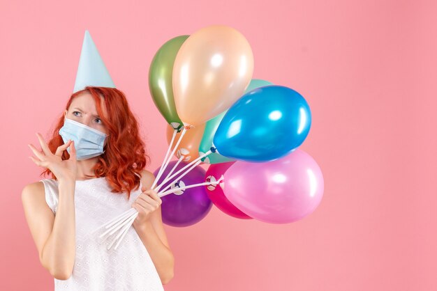 Vue de face de la jeune femme tenant des ballons colorés en masque stérile sur le mur rose