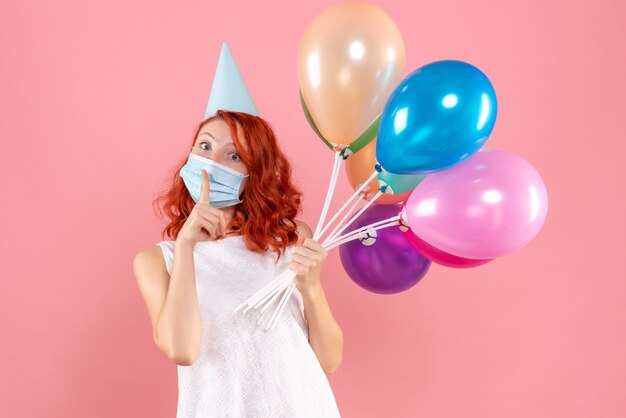 Vue de face de la jeune femme tenant des ballons colorés en masque stérile sur le mur rose