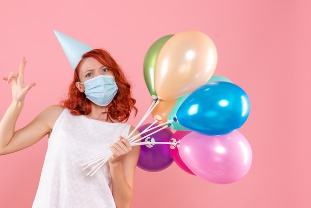 Vue de face de la jeune femme tenant des ballons colorés en masque stérile sur le mur rose