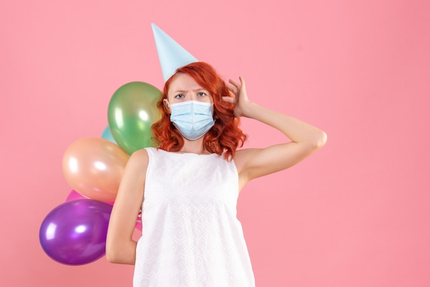 Vue de face de la jeune femme tenant des ballons colorés en masque stérile sur le mur rose