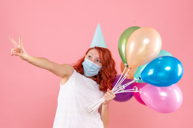 Vue de face de la jeune femme tenant des ballons colorés en masque stérile sur le mur rose