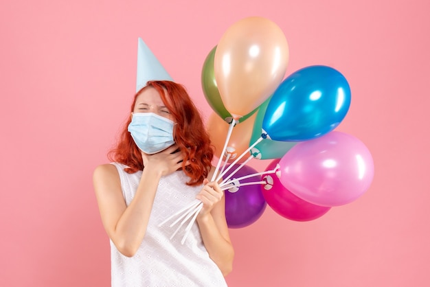 Vue de face de la jeune femme tenant des ballons colorés en masque stérile sur le mur rose