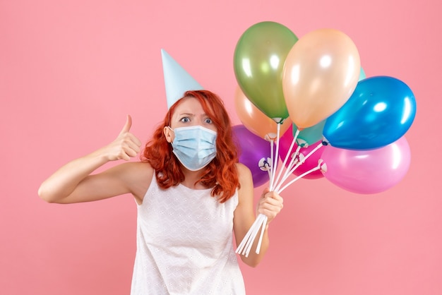 Vue de face jeune femme tenant des ballons colorés en masque stérile sur rose