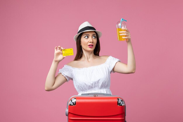 Vue de face jeune femme tenant une boisson et une carte bancaire sur un bureau rose voyage vacances été couleur chaleur