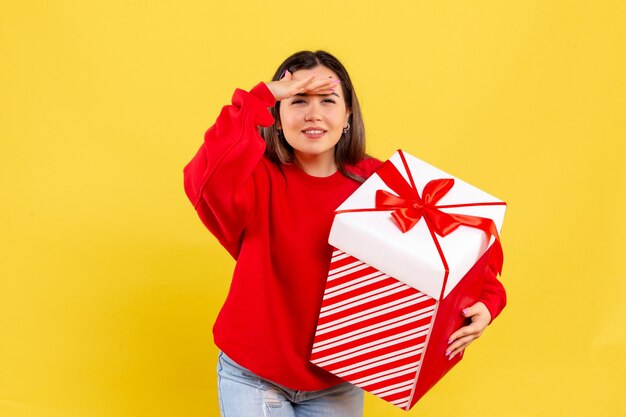 Vue de face de la jeune femme tenant un cadeau de Noël dans une boîte à la recherche sur le mur jaune