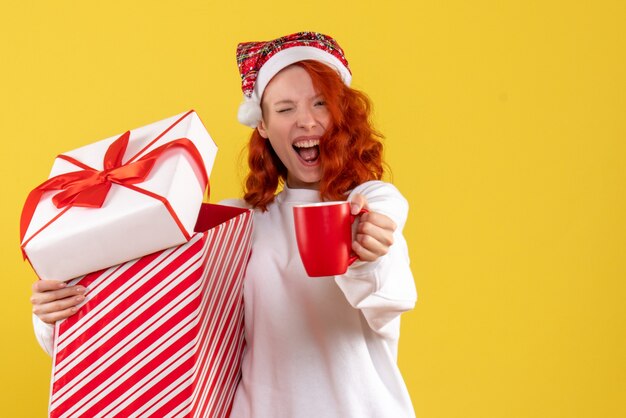 Vue de face de la jeune femme tenant un cadeau de Noël et une tasse de thé sur un mur jaune