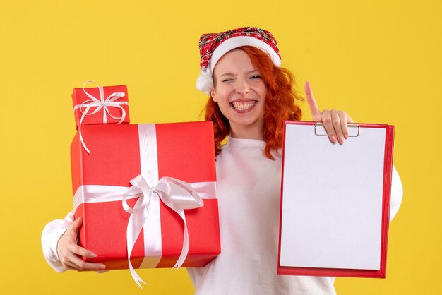 Vue de face de la jeune femme tenant des cadeaux de Noël et note de dossier sur le mur jaune