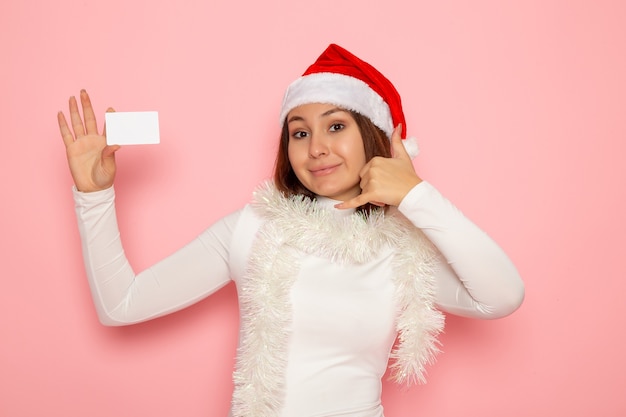 Vue de face jeune femme tenant une carte bancaire blanche sur le mur rose de Noël couleur vacances nouvel an