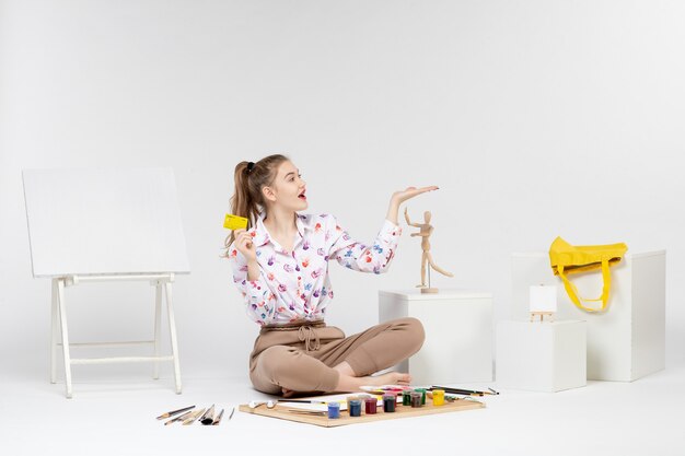 Vue de face jeune femme tenant une carte bancaire jaune sur un bureau blanc peintre chevalet artiste argent couleur dessiner art