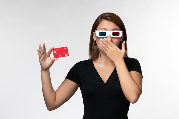 Vue de face jeune femme tenant une carte bancaire rouge dans des lunettes de soleil sur une surface blanche