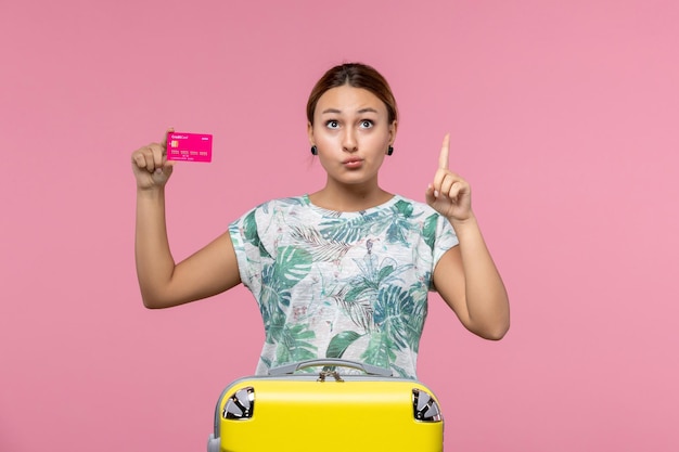 Vue de face jeune femme tenant une carte bancaire en vacances sur le mur rose couleurs de repos d'été voyage vacances femme