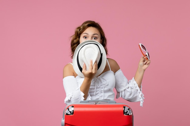 Vue de face d'une jeune femme tenant un chapeau et des lunettes de soleil avec un sac rouge sur le mur rose