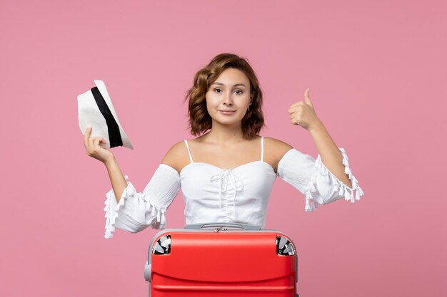 Vue de face d'une jeune femme tenant un chapeau et se préparant pour un voyage avec un sac rouge sur un mur rose clair