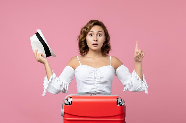 Vue de face d'une jeune femme tenant un chapeau et se préparant pour un voyage avec un sac rouge sur le mur rose