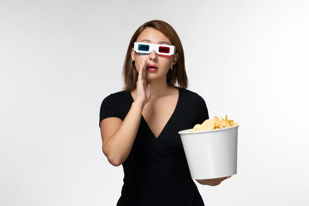 Vue de face jeune femme tenant des chips de pommes de terre dans des lunettes de soleil et regarder un film sur une surface blanche légère