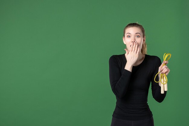 Vue de face jeune femme tenant une corde à sauter avec un visage surpris sur fond vert femme entraînement couleur sport corps gymnastique santé athlète