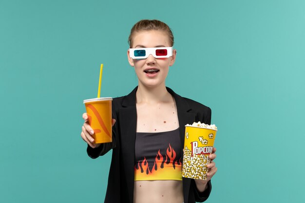 Vue de face jeune femme tenant du pop-corn avec boisson dans des lunettes de soleil et regarder un film sur un bureau bleu