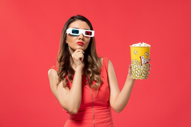 Vue de face jeune femme tenant du pop-corn dans des lunettes de soleil d pensant sur une surface rouge clair