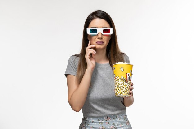 Vue de face jeune femme tenant du pop-corn et regarder un film dans des lunettes de soleil sur la surface blanche claire