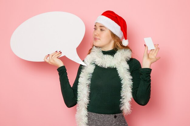 Vue de face jeune femme tenant grand panneau blanc et carte bancaire sur mur rose modèle vacances noël