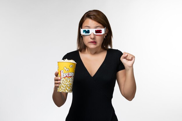 Vue de face jeune femme tenant un paquet de pop-corn dans des lunettes de soleil et regarder un film sur une surface blanche