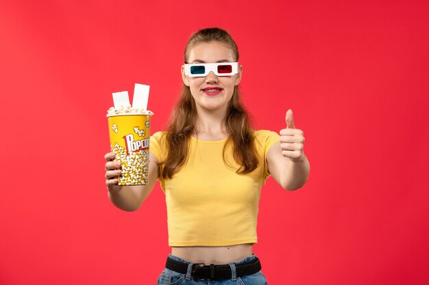 Vue de face jeune femme tenant le paquet de pop-corn en d lunettes de soleil sur mur rouge clair cinéma cinéma film cinéma