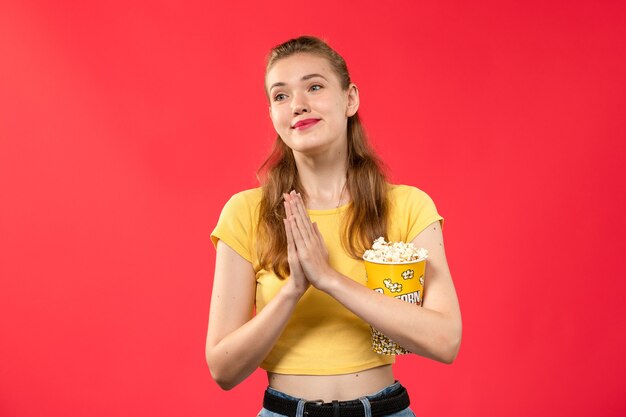 Vue de face jeune femme tenant le paquet de pop-corn et posant sur un mur rouge clair cinéma film cinéma film