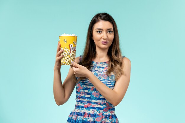 Vue de face jeune femme tenant le paquet de pop-corn sur la surface bleu clair