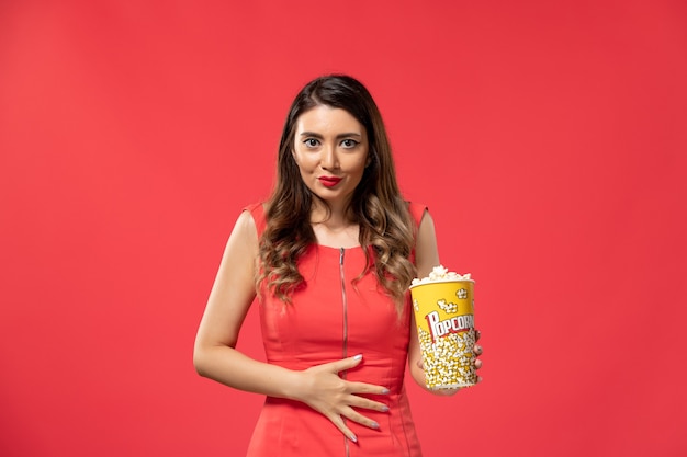 Vue de face jeune femme tenant le paquet de pop-corn sur la surface rouge clair