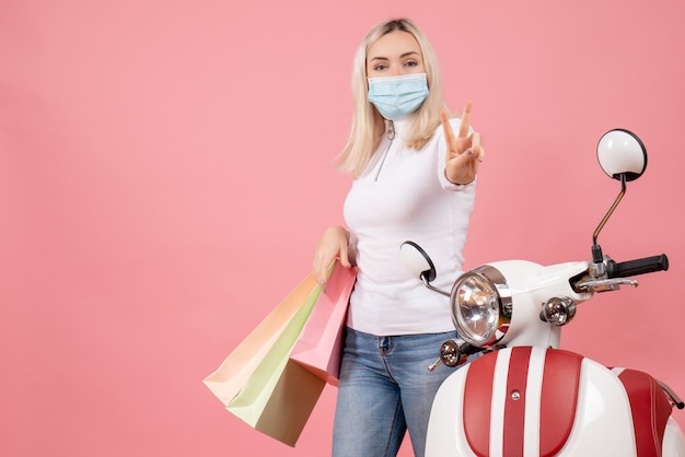 Vue de face jeune femme tenant des sacs à provisions gestes signe de la victoire près de cyclomoteur