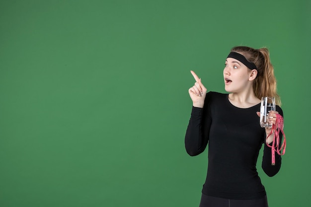 Vue de face jeune femme tenant un tour de taille et un verre d'eau sur fond vert sport couleur santé corps femme athlète entraînement noir