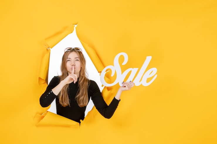 Photo gratuit vue de face jeune femme tenant une vente par écrit à travers un trou de papier déchiré dans le mur