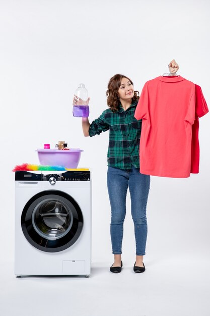 Vue de face d'une jeune femme tenant des vêtements propres et de la poudre liquide de la machine à laver sur un mur blanc