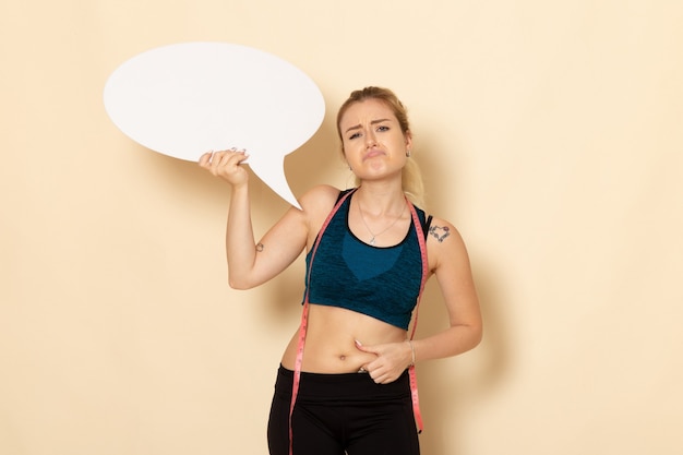 Vue de face jeune femme en tenue de sport tenant une pancarte blanche et mesurant son corps sur un mur blanc léger corps santé sport beauté exercices fit