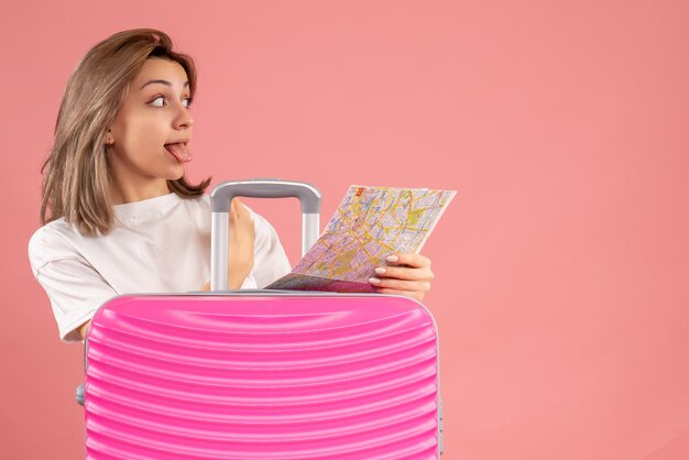 Vue de face jeune femme avec une valise rose qui sort la langue tenant la carte