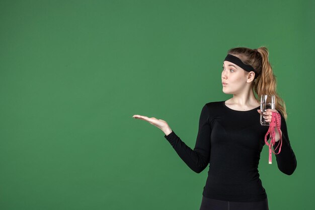 Vue de face jeune femme avec verre d'eau et mesure de la taille sur fond vert couleurs santé corps noir entraînement femme athlète sport