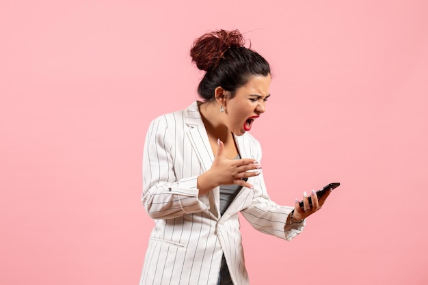 Vue de face jeune femme en veste blanche tenant un téléphone et lui criant dessus sur fond rose émotion femme mode sentiment couleur dame